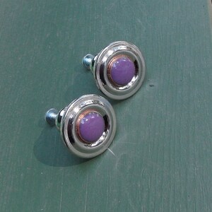 Peut inclure: Deux boutons de meuble argent&eacute;s avec un design circulaire. Chaque bouton pr&eacute;sente un centre lavande, un anneau cuivr&eacute; et un anneau ext&eacute;rieur argent&eacute;. Les boutons ont une vis de fixation et sont pos&eacute;s sur une surface verte.