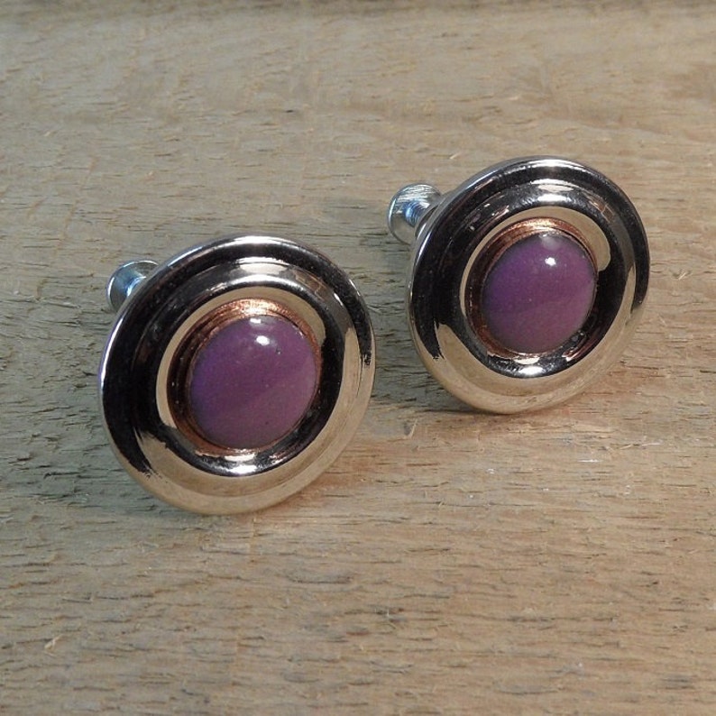 Peut inclure: Deux boutons de manchette argent&eacute;s de forme circulaire. Chaque bouton de manchette pr&eacute;sente une pierre centrale ronde de couleur violette, entour&eacute;e d'un anneau cuivr&eacute; et d'une bordure argent&eacute;e polie. Pr&eacute;sent&eacute;s sur une surface en bois.