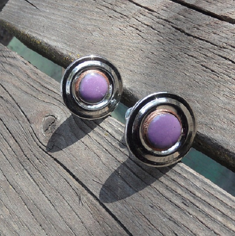 Peut inclure: Une paire de boucles d'oreilles rondes avec un design en m&eacute;tal argent&eacute;. Chaque boucle d'oreille pr&eacute;sente une pierre centrale circulaire, violet clair, entour&eacute;e d'anneaux concentriques et d'une bande couleur cuivre. Les boucles sont pos&eacute;es sur une surface en bois vieilli.