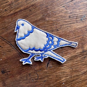 May include: Embroidered bird patch featuring a dove design. The bird has a light beige body with blue scalloped wing and tail details. The patch is outlined in blue and has blue feet and a beak.