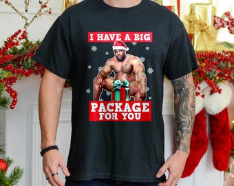 May include: A black t-shirt with a Christmas-themed design featuring a muscular man wearing a Santa hat and holding a gift box. The text on the shirt reads "I have a big package for you."