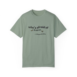 May include: Sage green t-shirt with the text "who's afraid of lil old me...well you should be" printed on the front. Two small butterfly graphics are above the text.
