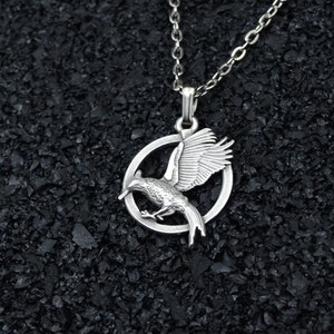 May include: Silver-toned necklace featuring a pendant with a bird design inside a circle. The bird has outstretched wings, and the chain is a delicate link style. The background is a dark, textured surface.