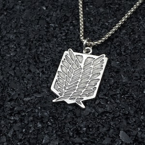 Könnte beinhalten: Eine silberne Halskette mit einem Anhänger in Form des Flügels der Freiheit Emblems aus dem Anime Attack on Titan. Der Anhänger ist mit einem strukturierten Design verziert und hängt an einer silbernen Kette.