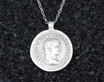 May include: A silver-colored pendant necklace featuring a circular coin design. The coin depicts a profile of a person with a laurel wreath and text around the edge. The necklace chain is also silver.