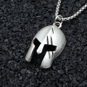 May include: A silver-toned necklace with a Spartan helmet pendant. The helmet is detailed with lines and a raised design.