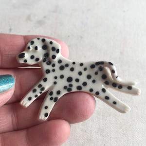 Op de afbeelding: Een keramiek dalmatiërhondje met zwarte vlekken op een witte achtergrond. Het beeldje staat in een rennende houding met zijn staart omhoog gekruld.
