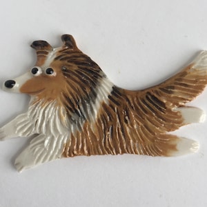 May include: A ceramic figurine of a brown and white Collie dog with black markings. The dog is depicted in a running pose with its tongue sticking out.