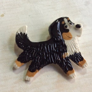 May include: A ceramic figurine of a Bernese Mountain Dog, painted in black, white, and brown. The dog is running with its tongue out and has a playful expression.