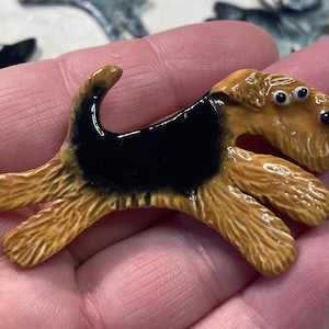 Op de afbeelding: Een keramiek beeldje van een bruin en zwarte draadhaar terriër met goggle-ogen. De hond staat in een rennende pose met zijn tong naar buiten.