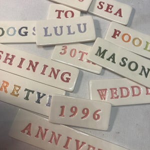 May include: A collection of rectangular white ceramic tiles featuring embossed letters and numbers in various colors. The tiles display text including 2026, SEA, TO, DOGS, LULU, FOOL, MASON, 30TH, SHINING, WEDD, RETY, 1996, and ANNIVERSARY.