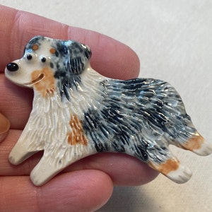 Può includere: Una statuetta in ceramica di un cane pastore australiano. Il cane è dipinto in tonalità di bianco, nero e marrone, con una texture di pelliccia dettagliata. La statuetta è in una posa di corsa, con la testa girata di lato.