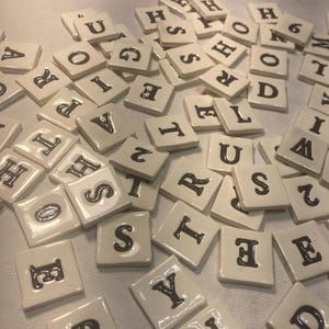 May include: A collection of square, off-white ceramic tiles, each featuring a single black letter or number printed in a serif font. The tiles are scattered in a random arrangement, showing various characters including letters like S, T, R, U, E, and numbers.