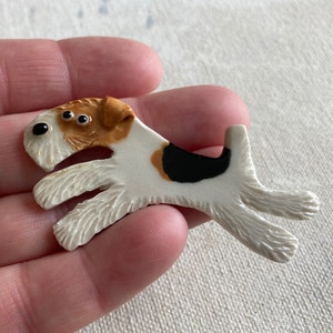 May include: A white, brown and black ceramic figurine of a wirehaired fox terrier dog. The figurine has a detailed face with black eyes and a brown nose.