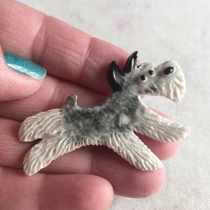 Op de afbeelding: Een kleine hondenfiguur in een rennende pose. De hond heeft een witte, getextureerde body, grijze markeringen en zwarte oren. Het beeldje is waarschijnlijk gemaakt van keramiek of een vergelijkbaar materiaal.