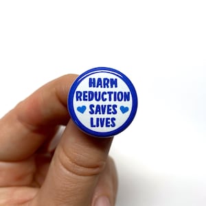 Puede incluir: Un bot&oacute;n azul y blanco con el texto "Harm Reduction Saves Lives" y dos corazones azules.