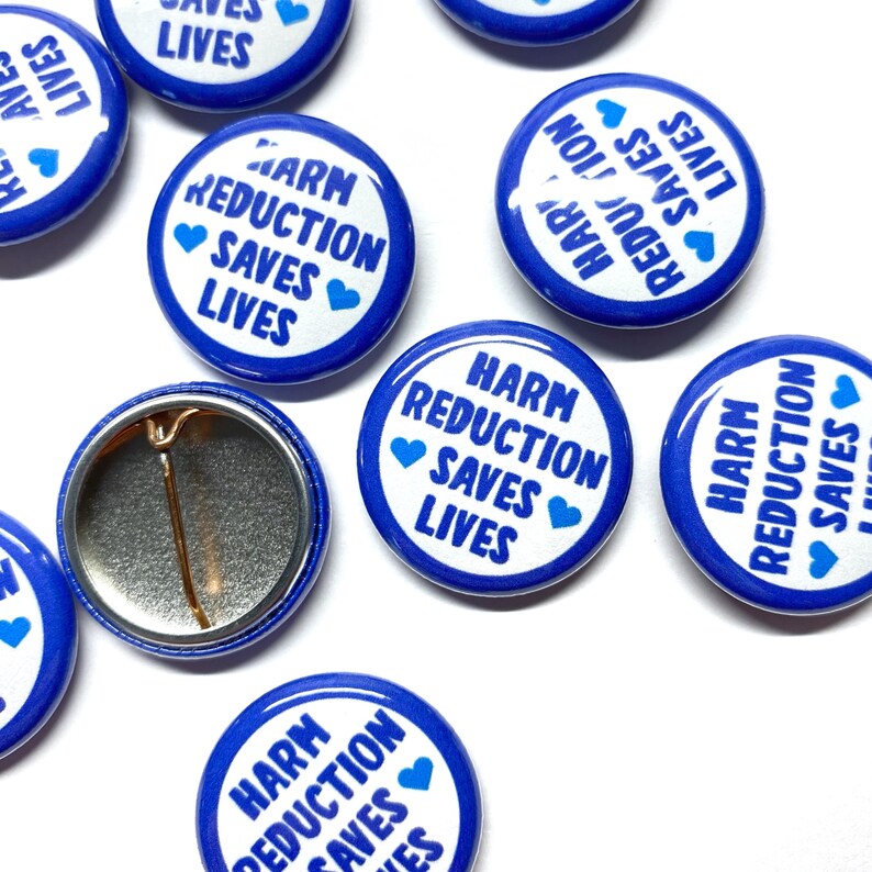 Puede incluir: Una colecci&oacute;n de botones azules y blancos con el texto "Harm Reduction Saves Lives" y un peque&ntilde;o coraz&oacute;n azul en cada bot&oacute;n.