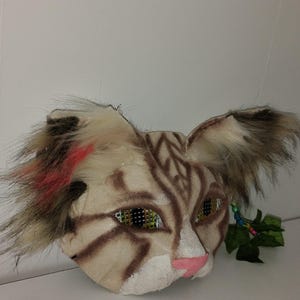 May include: A handmade cat mask with a tan and brown color scheme. The mask features faux fur accents, including tufts of fur on the ears and around the face. The eyes are green and the nose is pink.