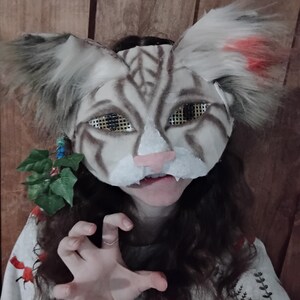 Op de afbeelding: Een kattenmasker met grote, pluizige oren en een gedetailleerd gezicht. Het masker heeft een witte basis met bruine strepen, een roze neus en mesh oogbedekkingen. De oren hebben grijze en rode accenten. Het masker maakt deel uit van een kostuum.