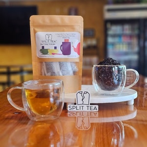 May include: A brown paper bag with a label that reads "SPLIT TEA" Earl Grey Lavender. The bag contains two tea bags. There are two glass mugs with tea, one with Earl Grey Lavender tea and the other with a dark tea. The mugs are on a white coaster. There is a sticker on the table that reads "SPLIT TEA".