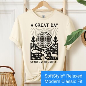 Cream color t-shirt featuring a folk art style graphic of a round waffle rising like a sun above a forest scene with woodland animals, plants, and a stream. Text above reads “A GREAT DAY” and below reads “STARTS WITH WAFFLES.”