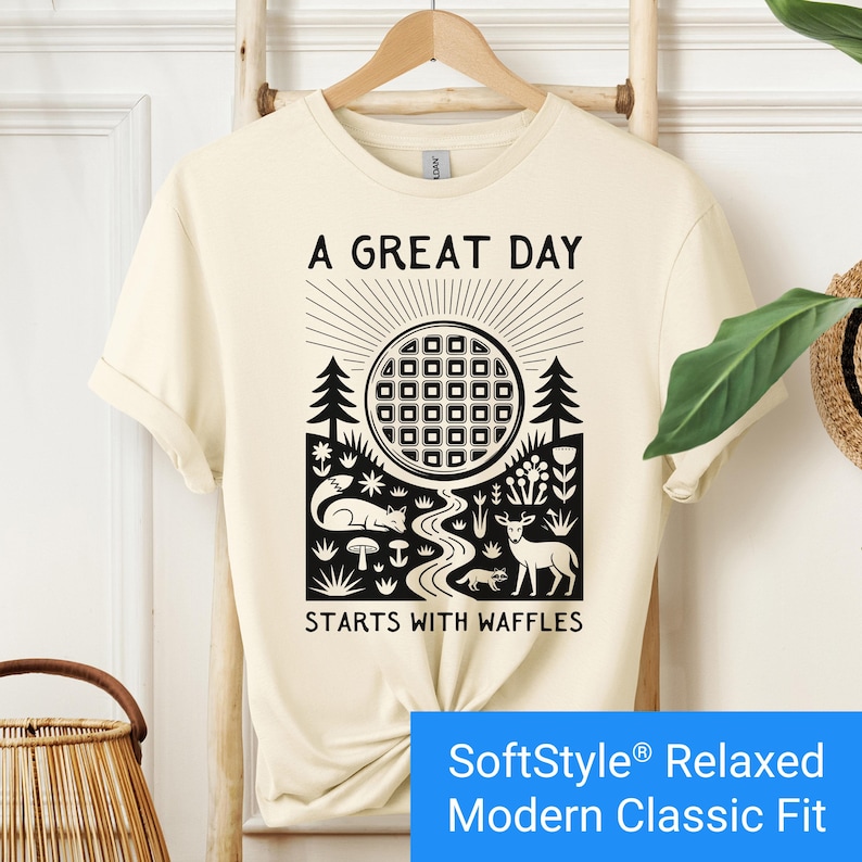 Cream color t-shirt featuring a folk art style graphic of a round waffle rising like a sun above a forest scene with woodland animals, plants, and a stream. Text above reads “A GREAT DAY” and below reads “STARTS WITH WAFFLES.”