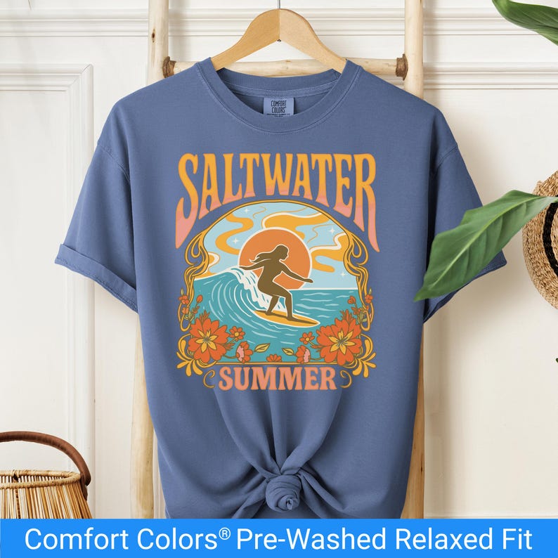 Blue jean color t-shirt featuring a retro-style design of a female surfer riding a wave in front of an orange sunset, surrounded by floral and ocean motifs with the words "Saltwater Summer" in bold vintage fonts.
