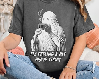 May include: Dark gray t-shirt featuring a white illustration of a ghost holding a coffee mug, with steam rising from it. The text "I'M FEELING A BIT GRAVE TODAY" is below the graphic. The shirt is a Comfort Colors Pre-Washed Relaxed Fit.