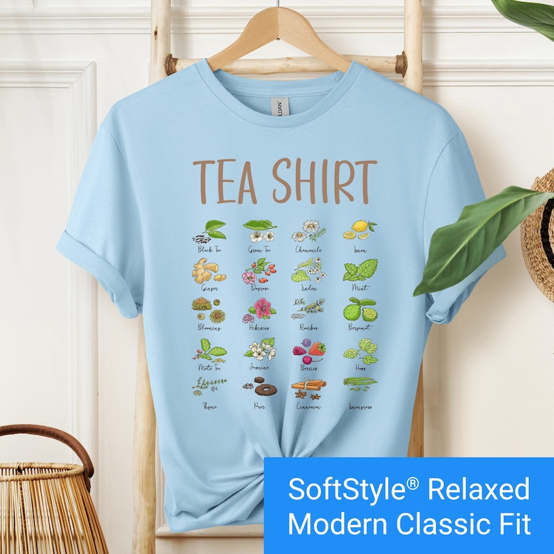Light blue t-shirt featuring a colorful illustrated grid of tea ingredients like mint, ginger, chamomile, and berries with large text that reads “Tea Shirt” at the top in brown lettering.