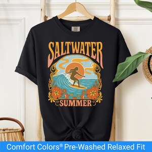 Black t-shirt featuring a retro-style design of a female surfer riding a wave in front of an orange sunset, surrounded by floral and ocean motifs with the words "Saltwater Summer" in bold vintage fonts.