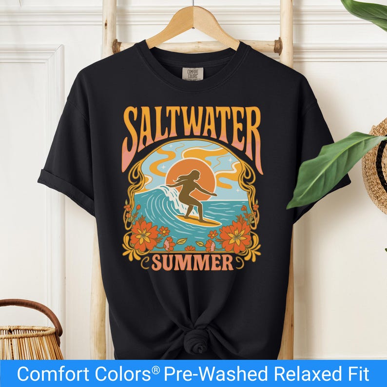 Black t-shirt featuring a retro-style design of a female surfer riding a wave in front of an orange sunset, surrounded by floral and ocean motifs with the words "Saltwater Summer" in bold vintage fonts.