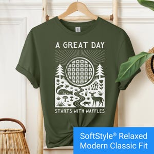 Military green t-shirt featuring a folk art style graphic of a round waffle rising like a sun above a forest scene with woodland animals, plants, and a stream. Text above reads “A GREAT DAY” and below reads “STARTS WITH WAFFLES.”