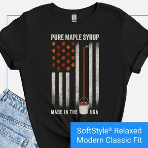 May include: Black t-shirt with a graphic design featuring a maple syrup-themed American flag. The design includes the text "PURE MAPLE SYRUP" and "MADE IN THE USA". The shirt is a SoftStyle Relaxed Modern Classic Fit.