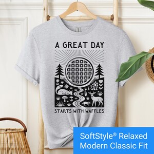 Sport grey t-shirt featuring a folk art style graphic of a round waffle rising like a sun above a forest scene with woodland animals, plants, and a stream. Text above reads “A GREAT DAY” and below reads “STARTS WITH WAFFLES.”