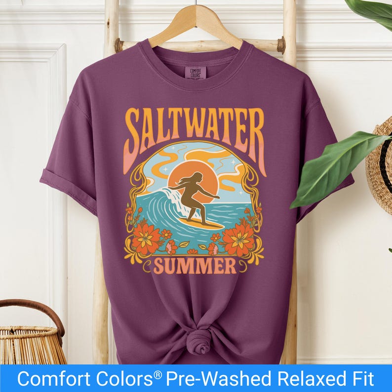 Berry color t-shirt featuring a retro-style design of a female surfer riding a wave in front of an orange sunset, surrounded by floral and ocean motifs with the words "Saltwater Summer" in bold vintage fonts.