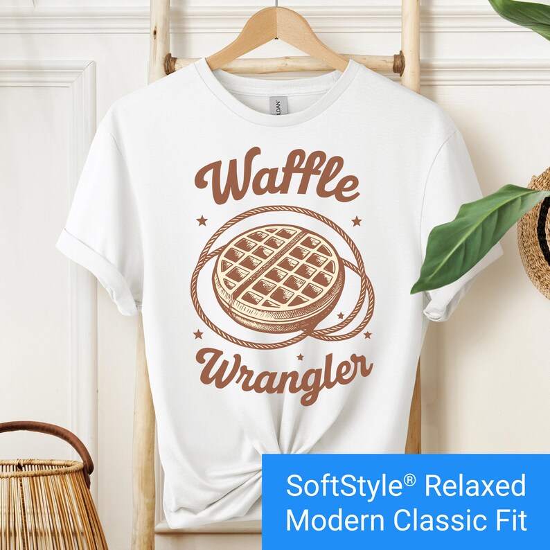 White t-shirt with a brown vintage-style graphic of a waffle encircled by a rope lasso, with the words "Waffle Wrangler" above and below the design.