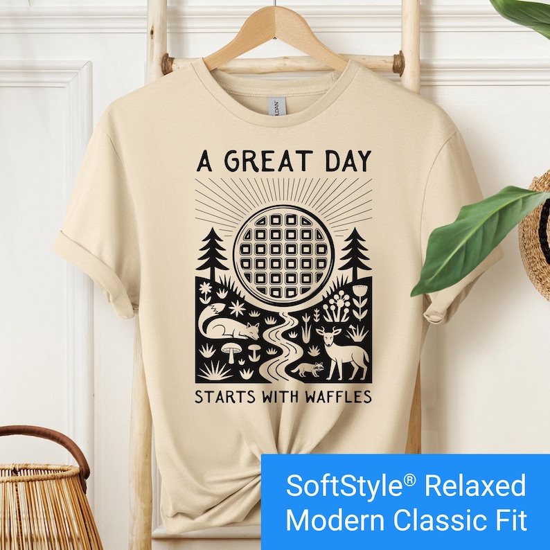 Sand color t-shirt featuring a folk art style graphic of a round waffle rising like a sun above a forest scene with woodland animals, plants, and a stream. Text above reads “A GREAT DAY” and below reads “STARTS WITH WAFFLES.”