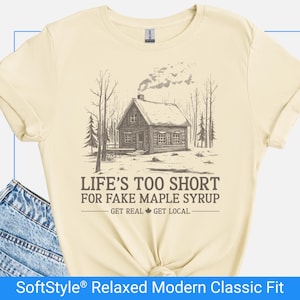 May include: Cream-colored t-shirt with a graphic of a cabin in a forest, smoke rising from the chimney. The text reads "LIFE'S TOO SHORT FOR FAKE MAPLE SYRUP - GET REAL GET LOCAL." The shirt is a SoftStyle Relaxed Modern Classic Fit.