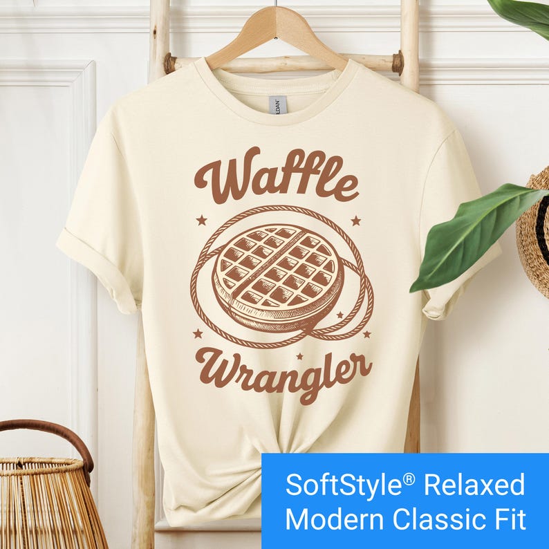 Cream color t-shirt with a brown vintage-style graphic of a waffle encircled by a rope lasso, with the words "Waffle Wrangler" above and below the design.