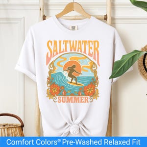 White t-shirt featuring a retro-style design of a female surfer riding a wave in front of an orange sunset, surrounded by floral and ocean motifs with the words "Saltwater Summer" in bold vintage fonts.