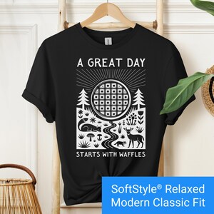 Black t-shirt featuring a folk art style graphic of a round waffle rising like a sun above a forest scene with woodland animals, plants, and a stream. Text above reads “A GREAT DAY” and below reads “STARTS WITH WAFFLES.”