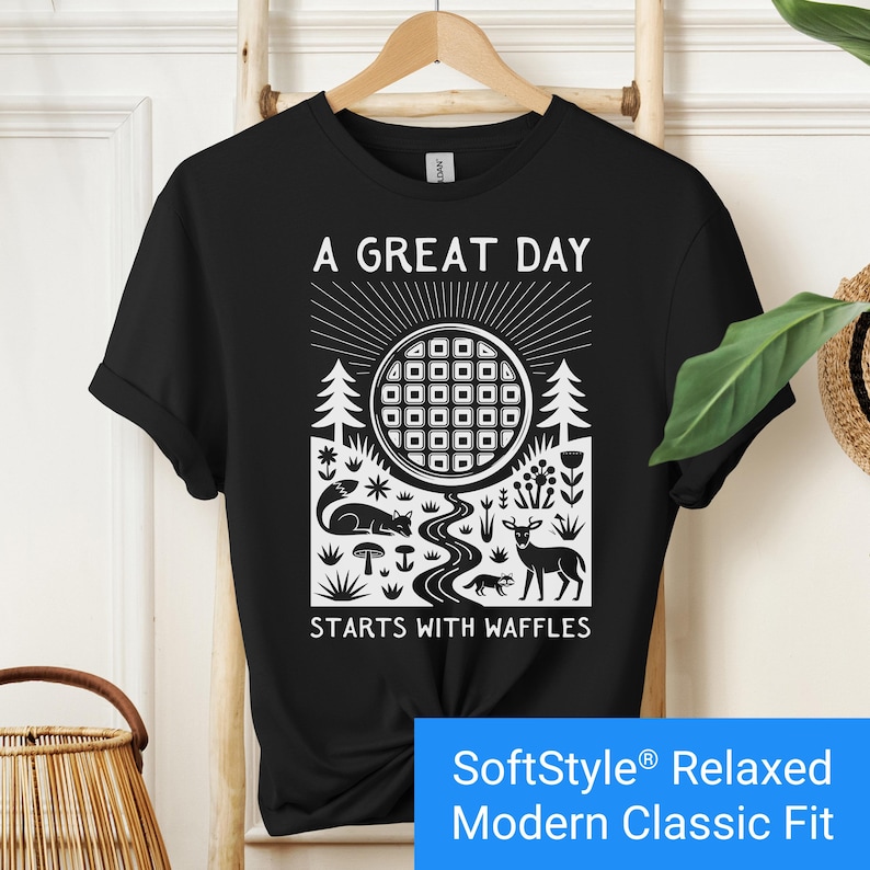 Black t-shirt featuring a folk art style graphic of a round waffle rising like a sun above a forest scene with woodland animals, plants, and a stream. Text above reads “A GREAT DAY” and below reads “STARTS WITH WAFFLES.”