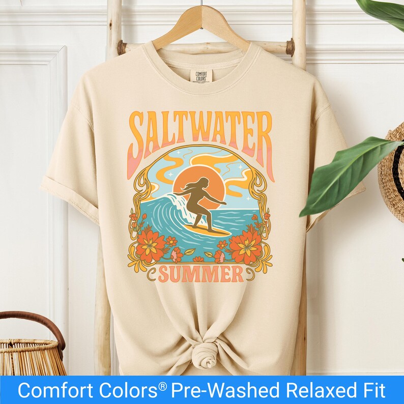 Ivory color t-shirt featuring a retro-style design of a female surfer riding a wave in front of an orange sunset, surrounded by floral and ocean motifs with the words "Saltwater Summer" in bold vintage fonts.
