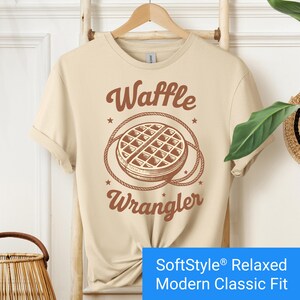 Sand color t-shirt with a brown vintage-style graphic of a waffle encircled by a rope lasso, with the words "Waffle Wrangler" above and below the design.