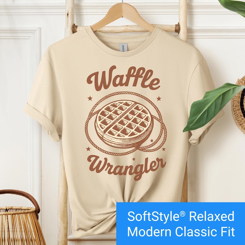 Sand color t-shirt with a brown vintage-style graphic of a waffle encircled by a rope lasso, with the words "Waffle Wrangler" above and below the design.