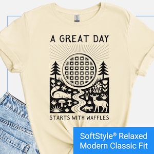 Cream color t-shirt featuring a folk art style graphic of a round waffle rising like a sun above a forest scene with woodland animals, plants, and a stream. Text above reads “A GREAT DAY” and below reads “STARTS WITH WAFFLES.”