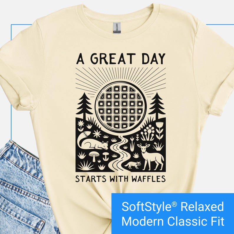 Cream color t-shirt featuring a folk art style graphic of a round waffle rising like a sun above a forest scene with woodland animals, plants, and a stream. Text above reads “A GREAT DAY” and below reads “STARTS WITH WAFFLES.”