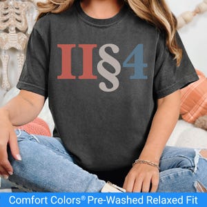 May include: A dark gray t-shirt with the text "II§4" in red, gray, and blue. The shirt is pre-washed and relaxed fit. The model is wearing blue jeans and a bracelet.