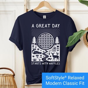 Navy blue t-shirt featuring a folk art style graphic of a round waffle rising like a sun above a forest scene with woodland animals, plants, and a stream. Text above reads “A GREAT DAY” and below reads “STARTS WITH WAFFLES.”