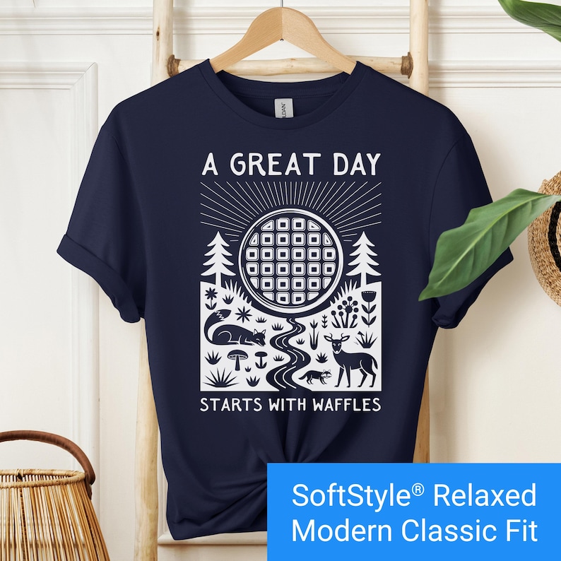 Navy blue t-shirt featuring a folk art style graphic of a round waffle rising like a sun above a forest scene with woodland animals, plants, and a stream. Text above reads “A GREAT DAY” and below reads “STARTS WITH WAFFLES.”